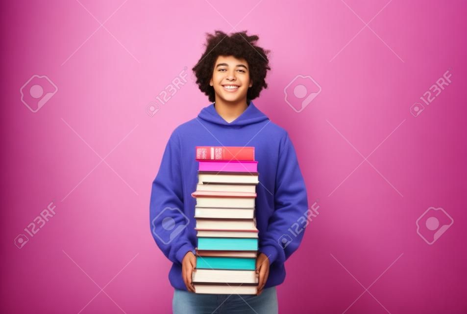 Adolescente allegro in piedi con una grande pila di libri, con un sacco di compiti su sfondo rosa per studio