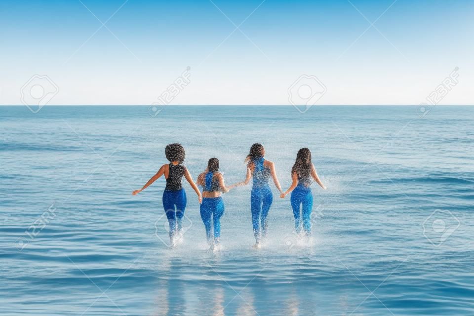 Back view of group of diverse female friends splashing water while running together at the beach, free space