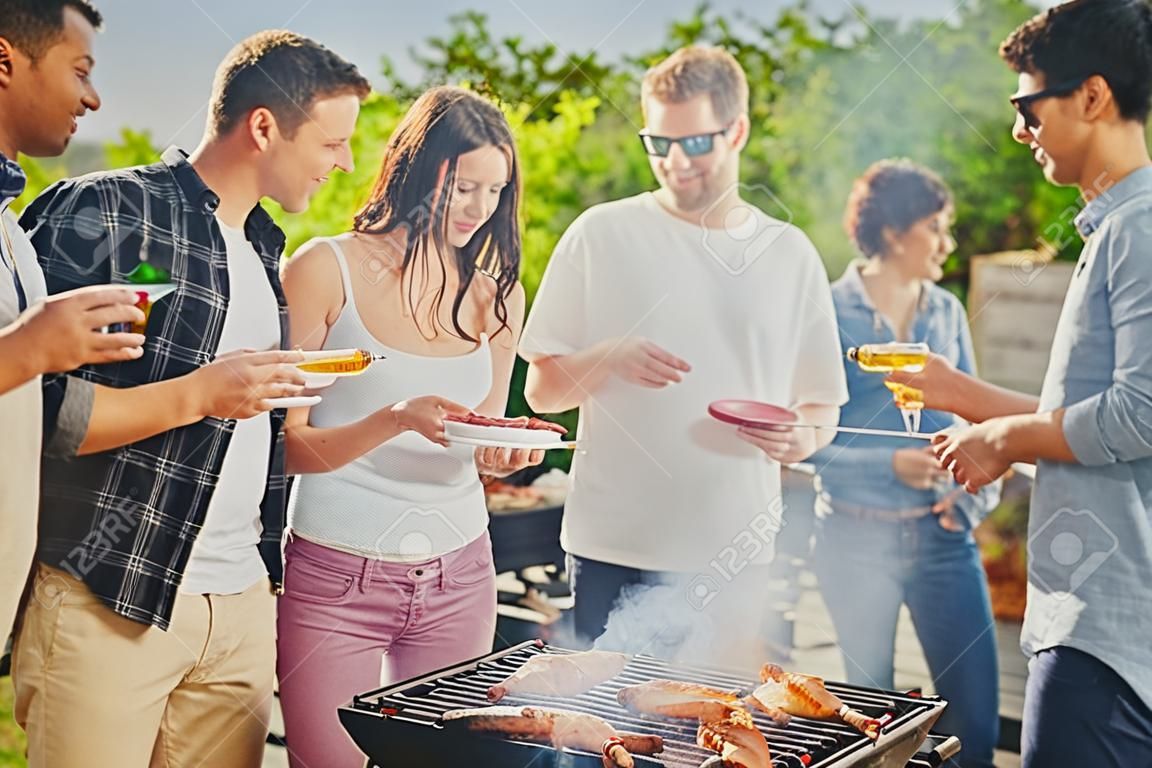 Group of people standing around grill, chatting, drinking and eating.