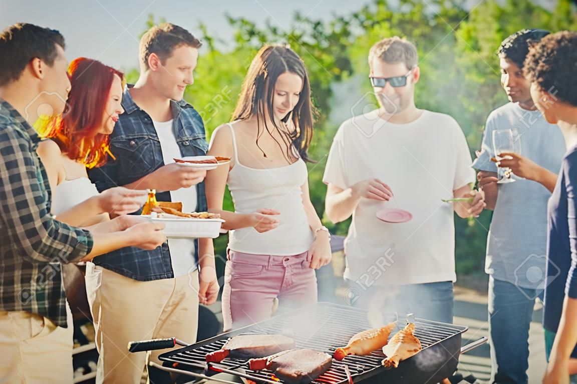 一群人站在烧烤，聊天，喝酒和吃东西。