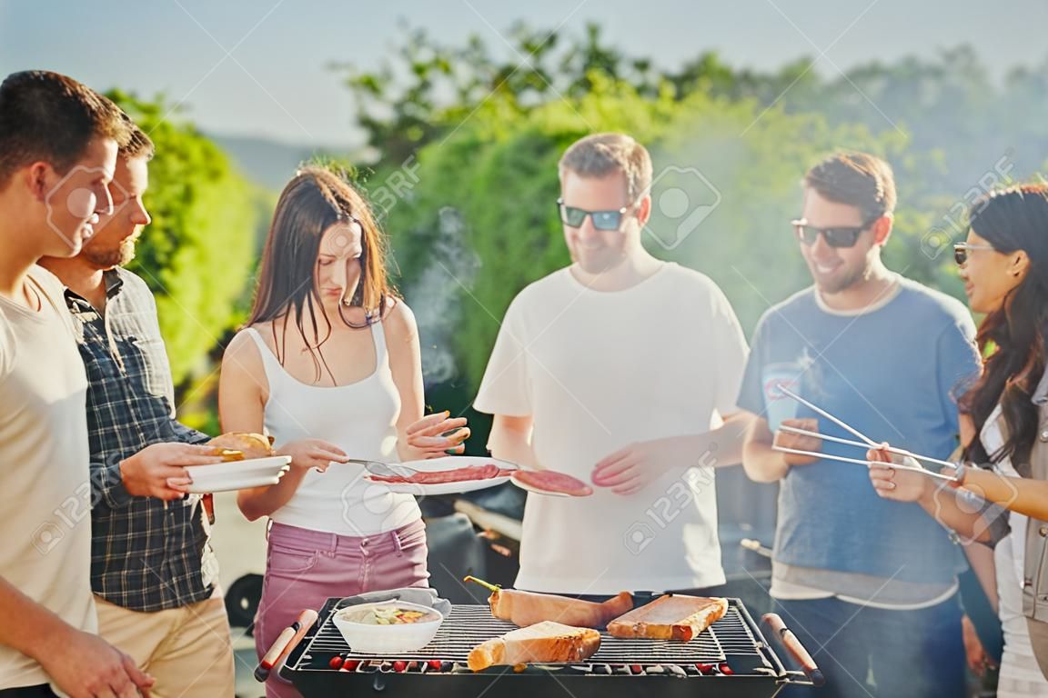 Group of people standing around grill, chatting, drinking and eating.