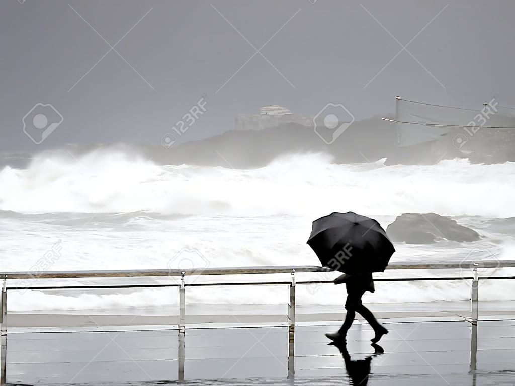 persona che protegge con l'ombrello in una giornata piovosa e ventosa che cammina sul lungomare con mare agitato