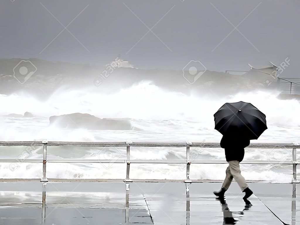 persona che protegge con l'ombrello in una giornata piovosa e ventosa che cammina sul lungomare con mare agitato