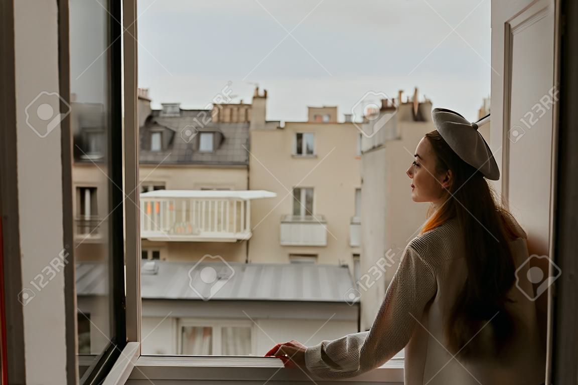 Une femme se tient près de la fenêtre sur fond de toits de Paris. Une fille aux cheveux longs en costume beige et béret profite de la ville et du voyage.