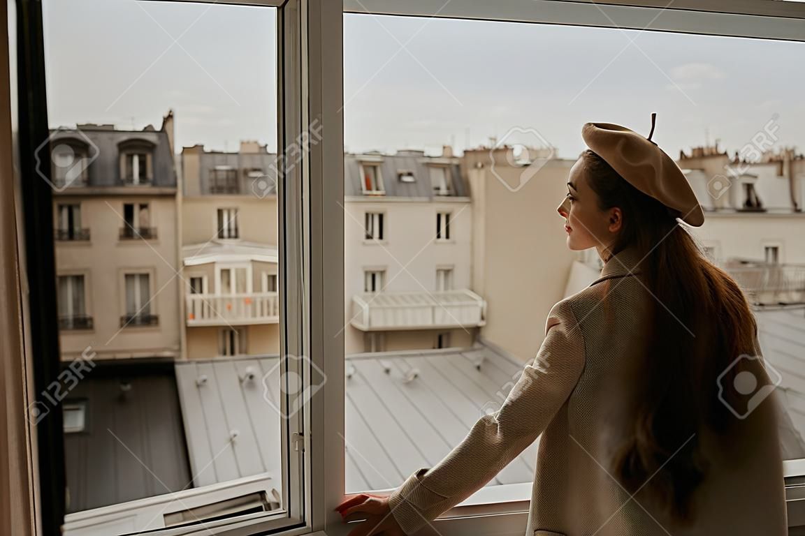 Une femme se tient près de la fenêtre sur fond de toits de Paris. Une fille aux cheveux longs en costume beige et béret profite de la ville et du voyage.