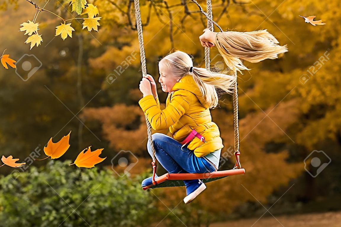 Szczęśliwa mała blondynka rasy kaukaskiej uśmiecha się i jeździ na huśtawce linowej jesienią w parku.