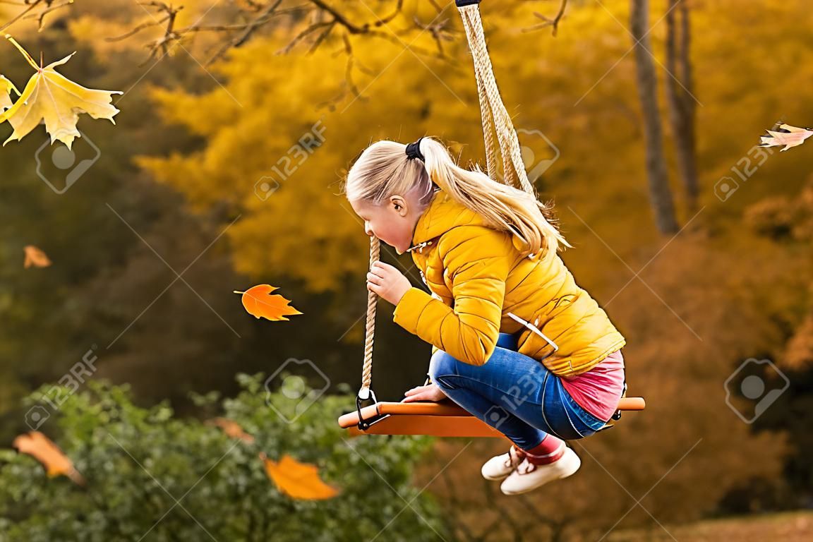 Szczęśliwa mała blondynka rasy kaukaskiej uśmiecha się i jeździ na huśtawce linowej jesienią w parku.