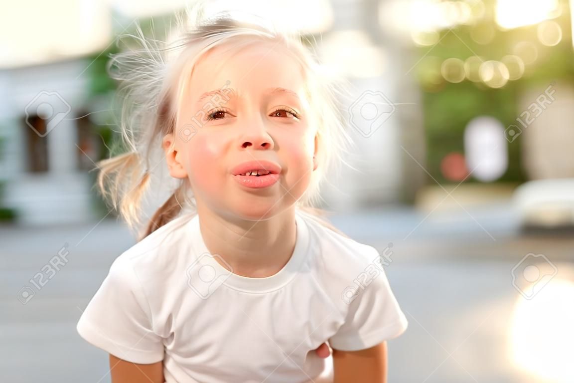 Cute smiling kid girl 4-5 year old showing tongue having fun on city street over sunset background outdoor. Looking at camera. Summer season. Childhood.