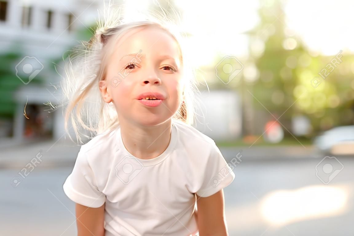 Cute smiling kid girl 4-5 year old showing tongue having fun on city street over sunset background outdoor. Looking at camera. Summer season. Childhood.
