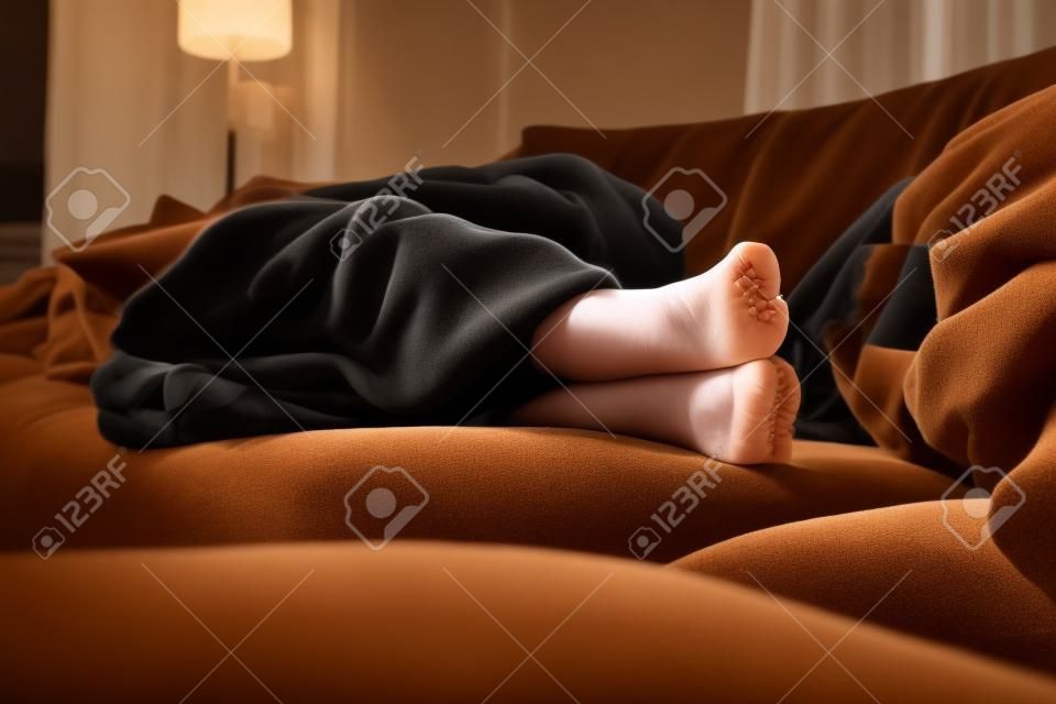 Ein Portrait von schwarzen Strumpfhosenfüßen eines Mädchens, während sie auf der Couch im Wohnzimmer unter einer braunen, kuscheligen Decke schläft.