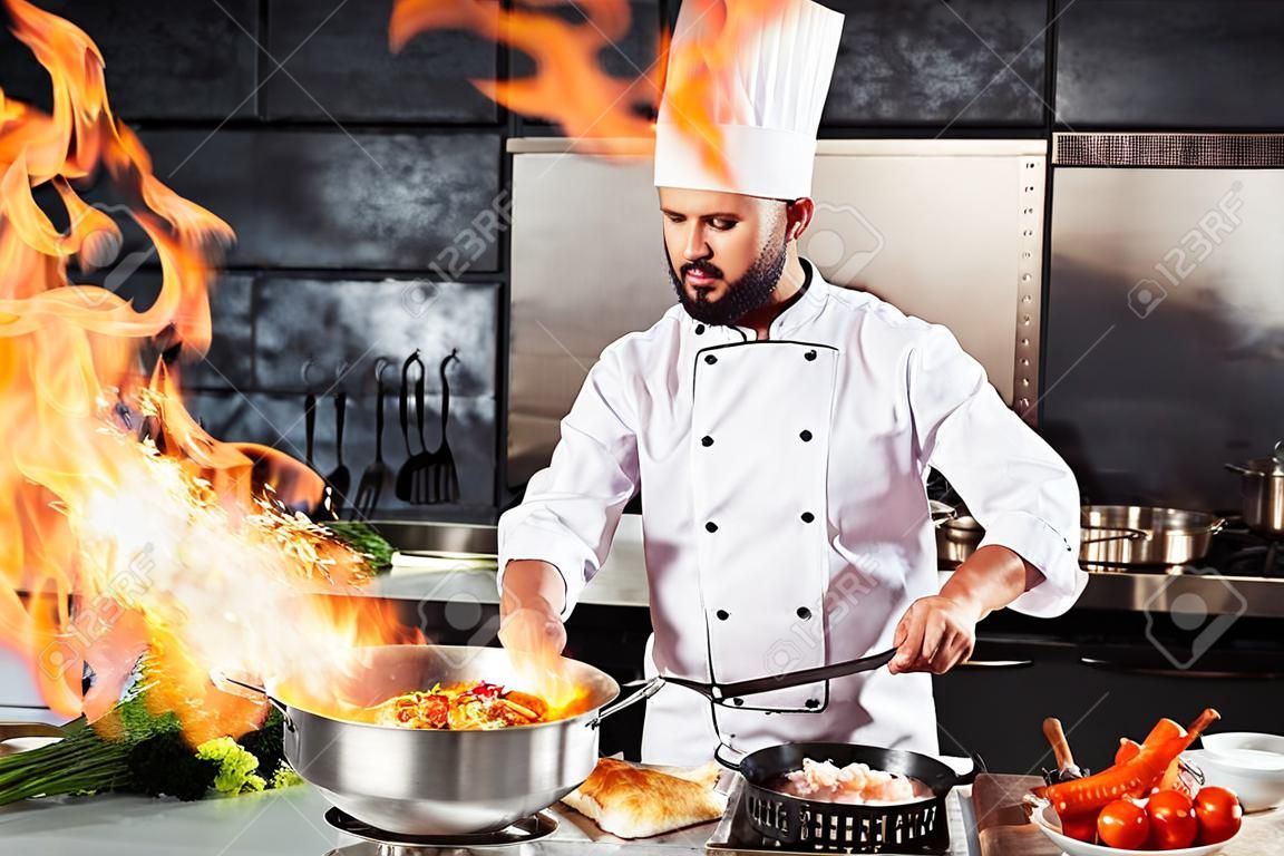 Chef cook food with fire at kitchen restaurant. Cook with wok at kitchen. Chef male in uniform hold wok with fire.