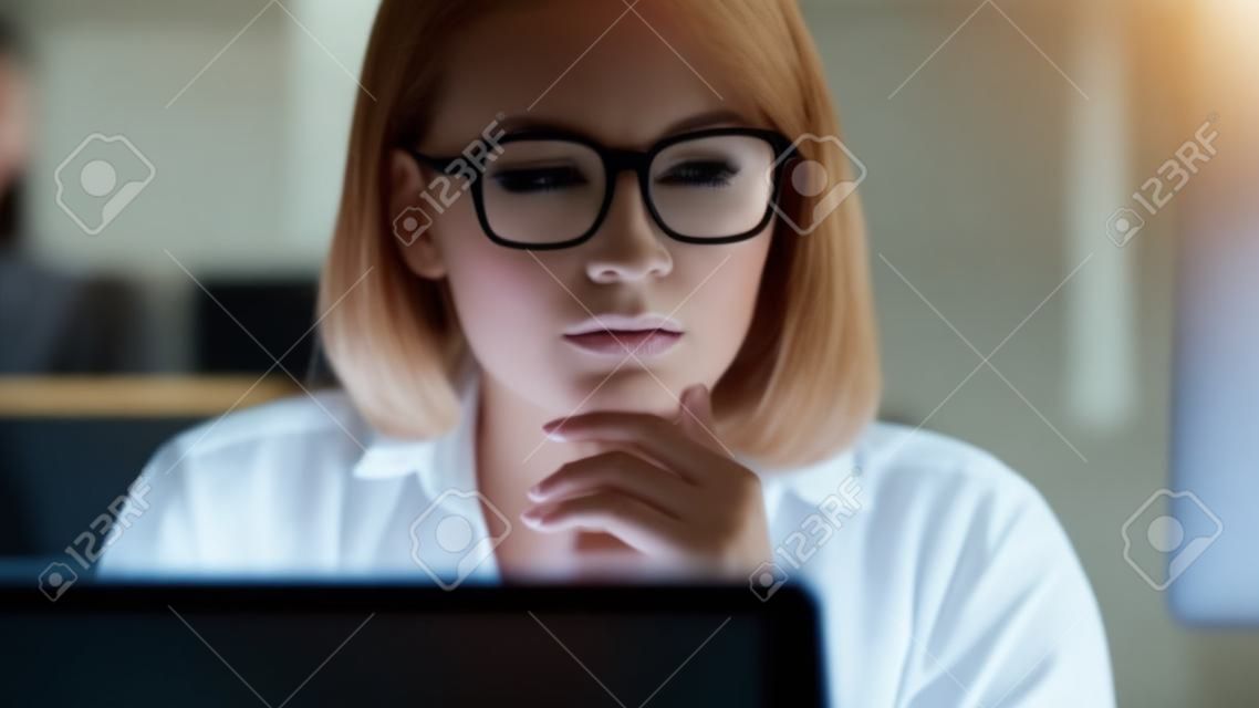Concentrated business woman looking laptop screen in dark office. Attentive woman in glasses watching financial report in computer monitor. Overworked business woman sitting in night office