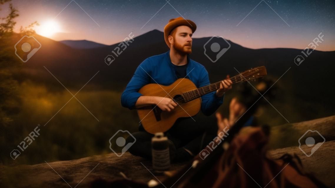 Il viaggiatore premuroso suona la chitarra all'aperto. ragazzo seduto vicino alle montagne natura falò da vicino. il ragazzo dai capelli rossi si gode la performance musicale sul paesaggio. il camper rilassato trascorre le vacanze la sera. concetto di voglia di viaggiare.