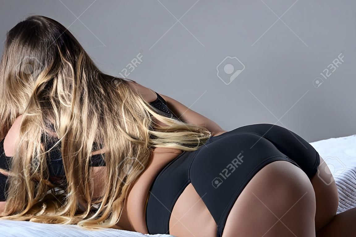 Na łóżku leży blondynka z długimi włosami w czarnej bieliźnie. Widok z tyłu dziewczyny w stringach śpiących na szarej kratce. Zgrabna kobieta z szerokimi biodrami, dużym tyłkiem i wąską talią. Zbliżenie.