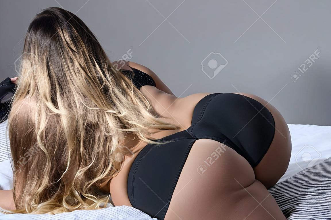 Uma loira com cabelo comprido em roupa interior preta está deitada na cama. Vista traseira de uma menina de tanga dormindo em um xadrez cinza. Uma mulher figurada com quadris largos, um grande espólio e uma cintura estreita. Close-up.