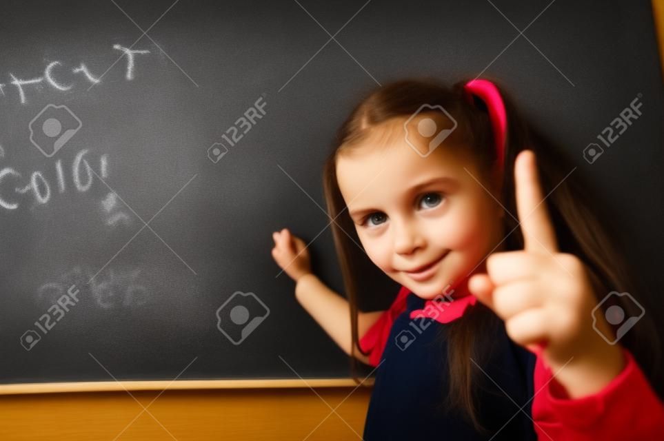 Portrait of a caucasian girl in the classroom. The schoolgirl writes the formula with chalk on the blackboard and looks into the camera.