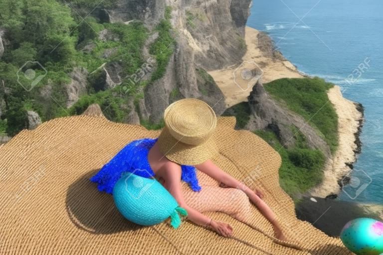 Straatfoto van een mooie vrouw met donker haar in een witte top, shorts en een hoed met een picknick op een heuvel met uitzicht op de zee