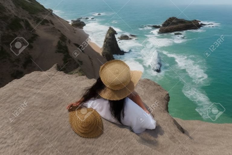 Straatfoto van een mooie vrouw met donker haar in een witte top, shorts en een hoed met een picknick op een heuvel met uitzicht op de zee