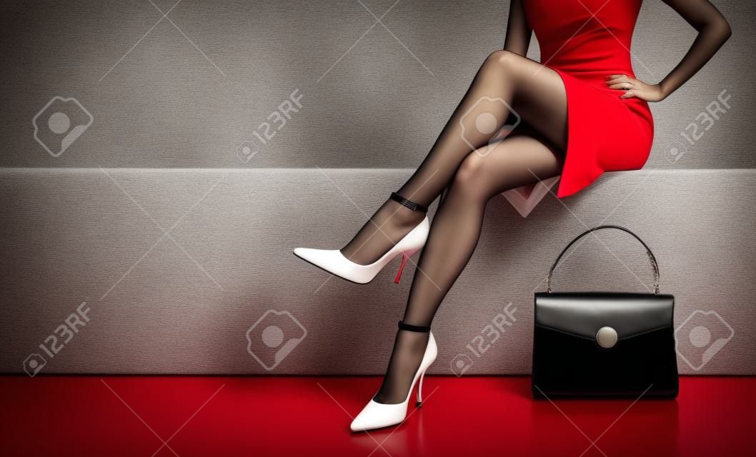 Piękne nogi kobieta nosi czerwoną sukienkę z czarnym portmonetka strony worka z Wysokie obcasy siedzi na białej ławce. z copyspace.
