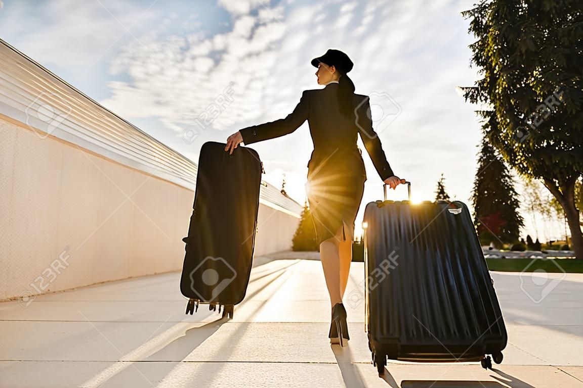 fiduciosa snella hostess donna in uniforme va su un volo con una valigia sotto i raggi del sole