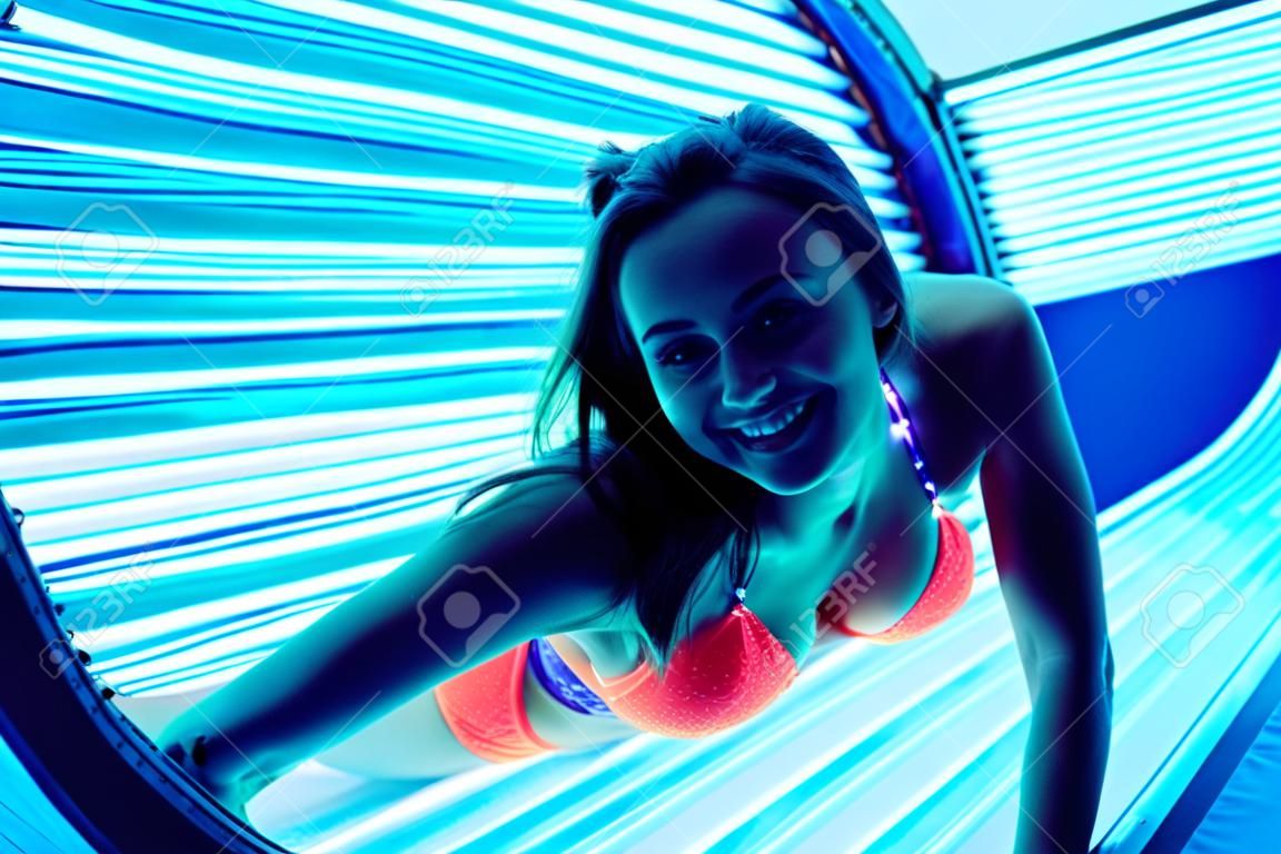 beautiful happy girl in swimsuit sunbathing in horizontal sunbed under ultraviolet rays, smiling