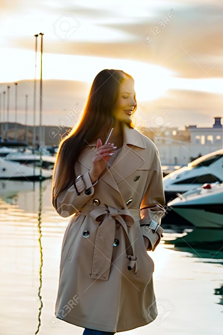 una lujosa chica de pelo largo con un moderno abrigo beige está esperando su yate en el puerto marítimo al atardecer