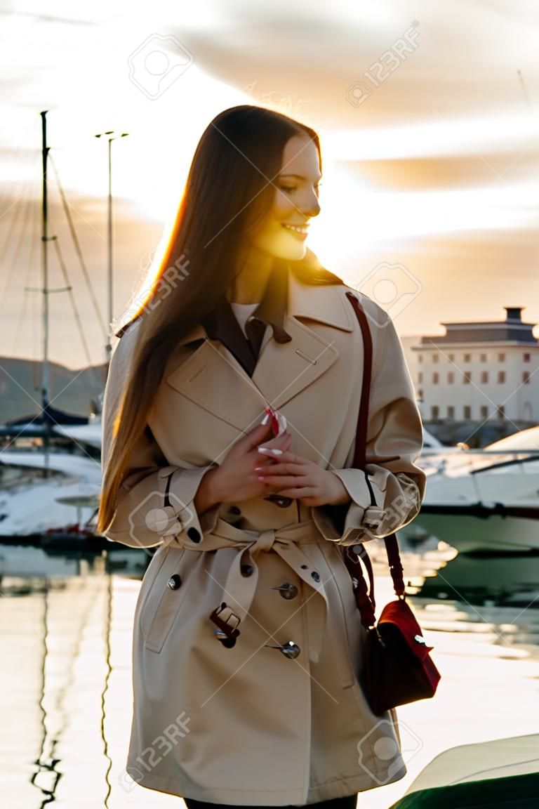 Luksusowa długowłosa dziewczyna w modnym beżowym płaszczu czeka na swój jacht w porcie morskim o zachodzie słońca