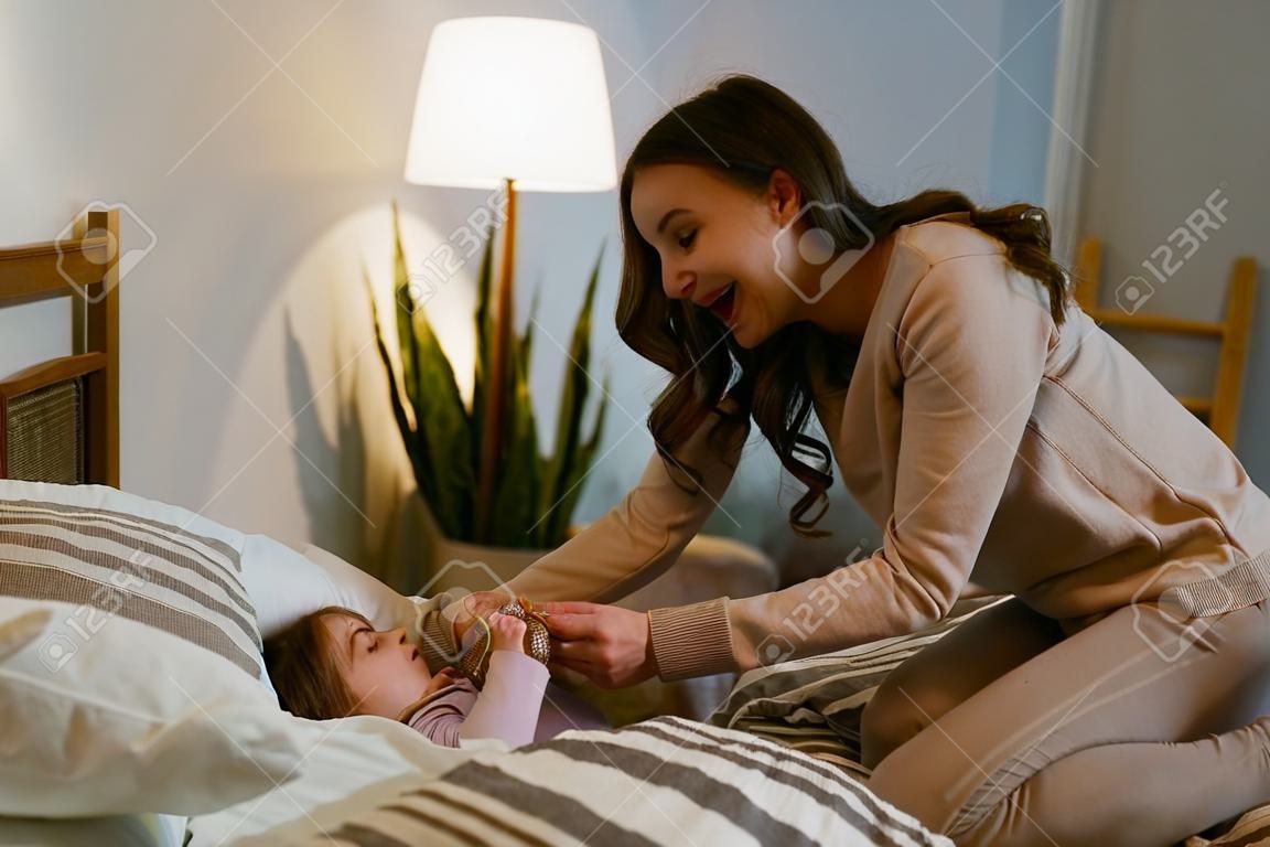 Cute mother in a beige sweater tickles and puts to bed a little daughter in the bedroom