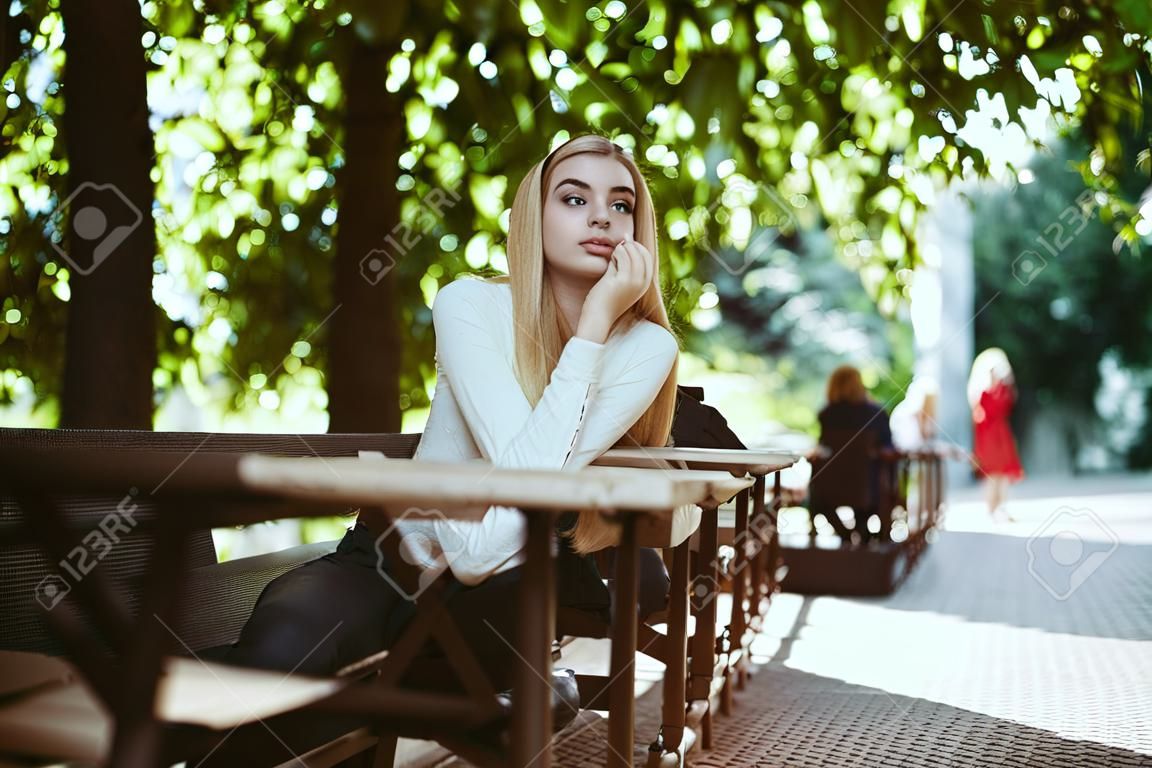 lovely blonde looks thoughtfully at the side resting her cheek on her hands in an outdoor cafe
