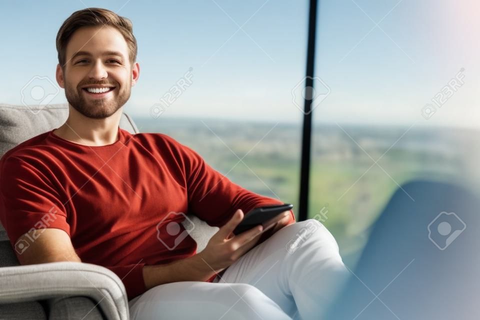 大きなガラス窓の横にあるモダンなソファに座っている白人男性は、携帯電話を手に持ったままカメラに向かって微笑んでいます。
