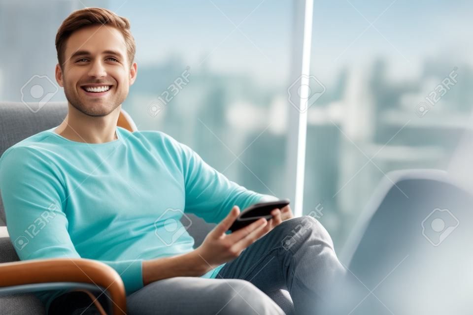 大きなガラス窓の横にあるモダンなソファに座っている白人男性は、携帯電話を手に持ったままカメラに向かって微笑んでいます。