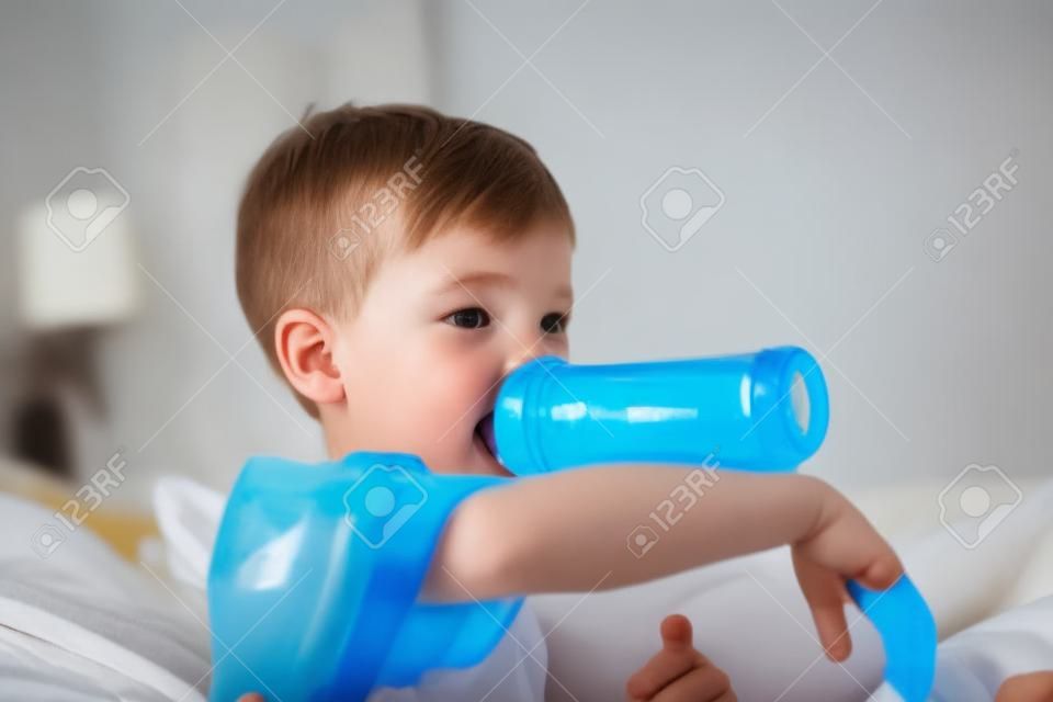 A boy is sucking a bottle of milk in the morning while watching TV in the bed in the bedroom.