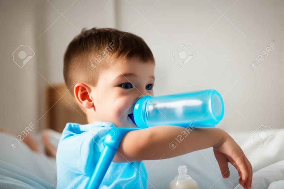 男の子は寝室のベッドでテレビを見ながら、午前中に牛乳のボトルを吸っています。
