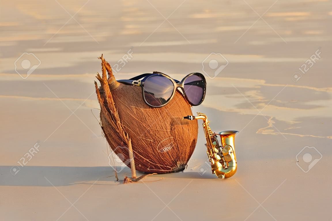 A coconut on the beach wearing sunglasses plays on a gold alto saxophone. Creative, humor and surrealism. Musical cover. Design with copy space.