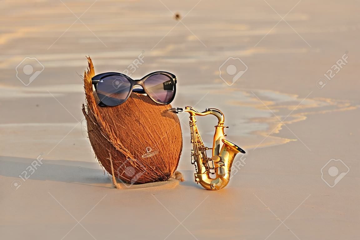 A coconut on the beach wearing sunglasses plays on a gold alto saxophone. Creative, humor and surrealism. Musical cover. Design with copy space.