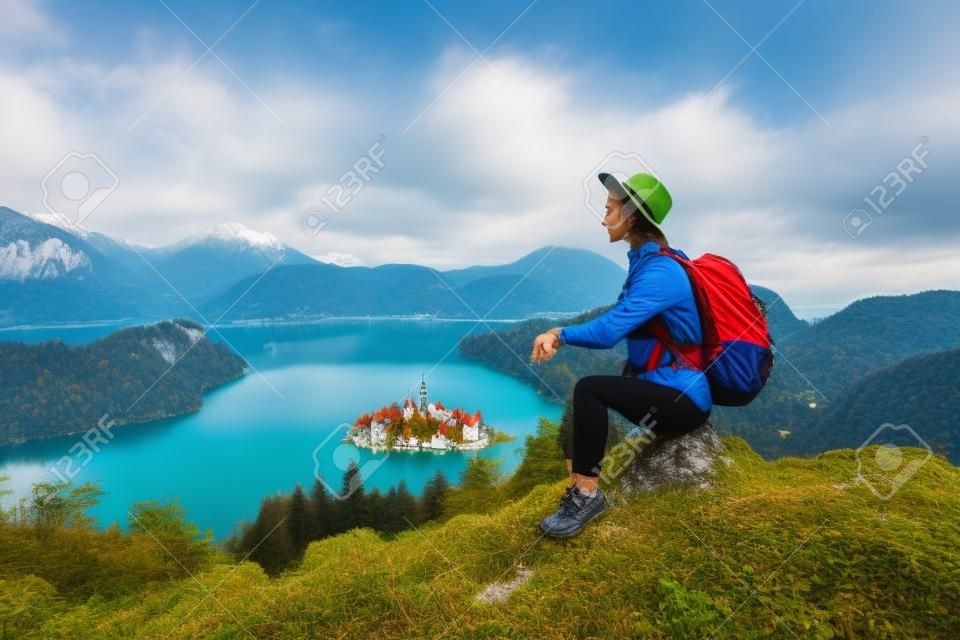 Travel Europe. Traveler woman in hat with backpack exploring Slovenia. Young hiker girl enjoying breathtaking view on amazing Bled Lake with Island and Alps Mountain. Autumn or winter nature landscape