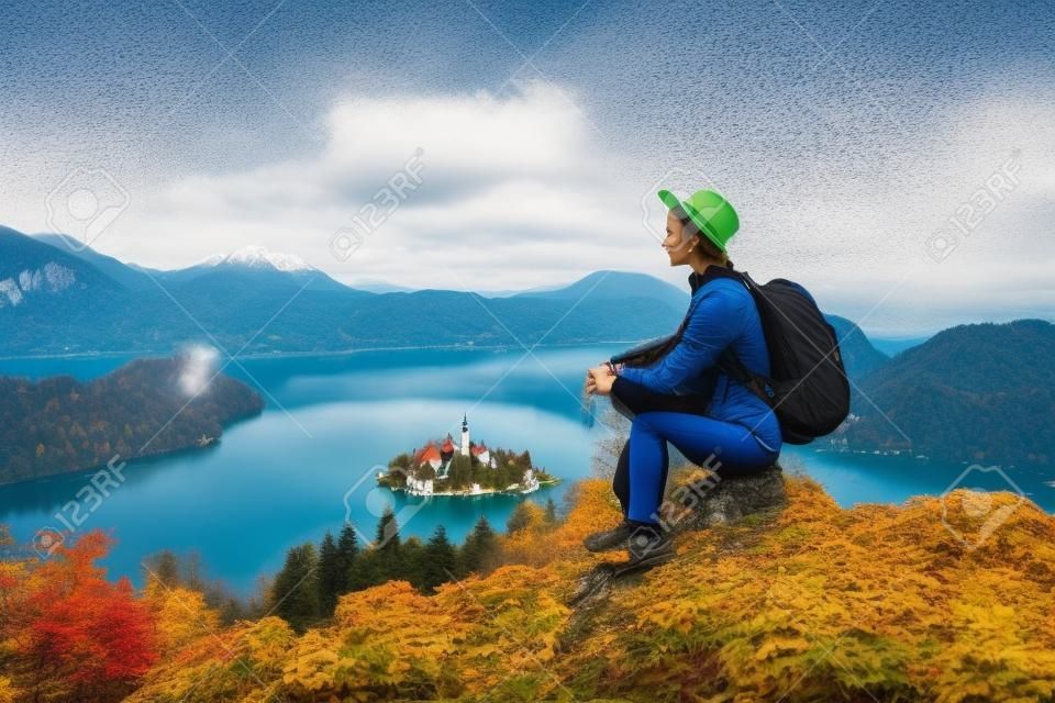 Travel Europe. Traveler woman in hat with backpack exploring Slovenia. Young hiker girl enjoying breathtaking view on amazing Bled Lake with Island and Alps Mountain. Autumn or winter nature landscape