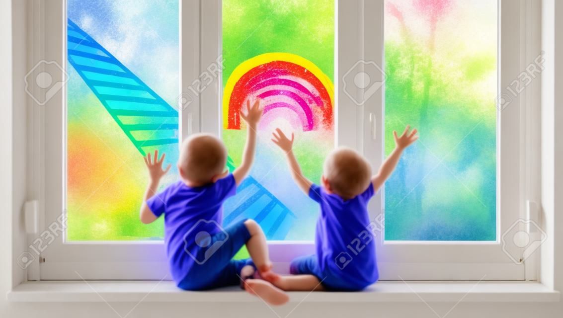 Małe dzieci na tle malarstwa tęczy na oknie. zdjęcie wypoczynku dzieci w domu. pozytywne wsparcie wizualne podczas kwarantanny pandemicznej koronawirusa covid-19 w domu. rodzinne tło