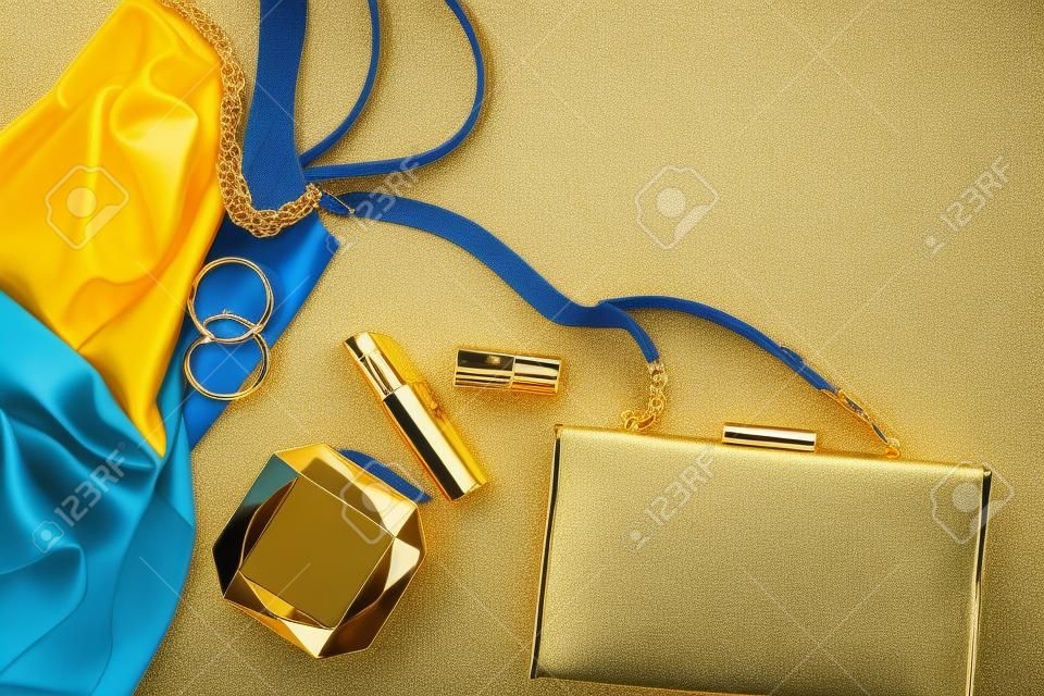 Women's gold-colored accessories cosmetics, handbag and cocktail dress in blue are on a light background. copyspace.