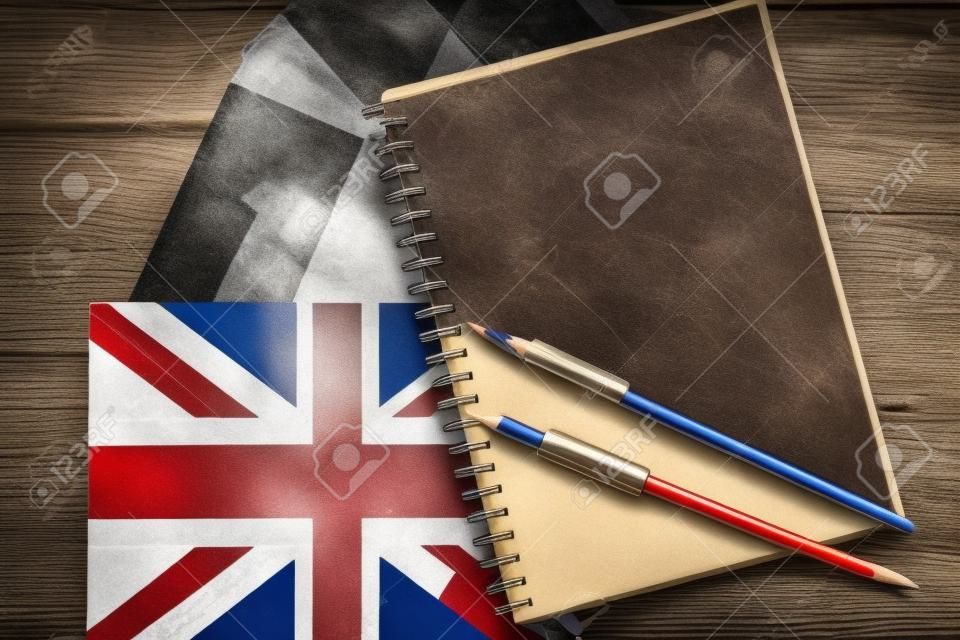 Three national flags, German, English, French, and notebook with blank sheets and two pencils on brown wooden surface. The concept of foreign language courses.