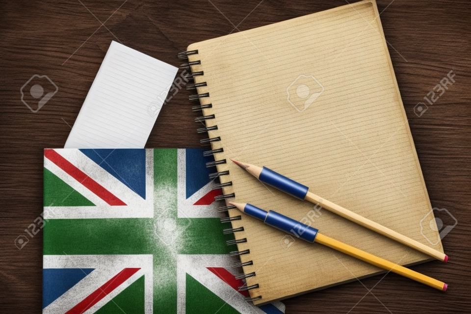 Three national flags, German, English, French, and notebook with blank sheets and two pencils on brown wooden surface. The concept of foreign language courses.