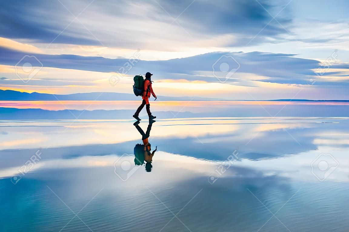 Mann Reisender mit Rucksack zu Fuß auf dem Salzsee bei Sonnenuntergang. Himmel mit Wolken spiegeln sich in der Spiegelwasseroberfläche wider. Reise- und Abenteuerkonzept