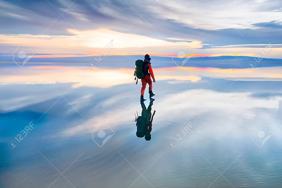 해가 질 때 배낭을 메고 소금 호수를 걷는 남자 여행자. 구름이 있는 하늘은 거울 수면에 반사됩니다. 여행과 모험 개념