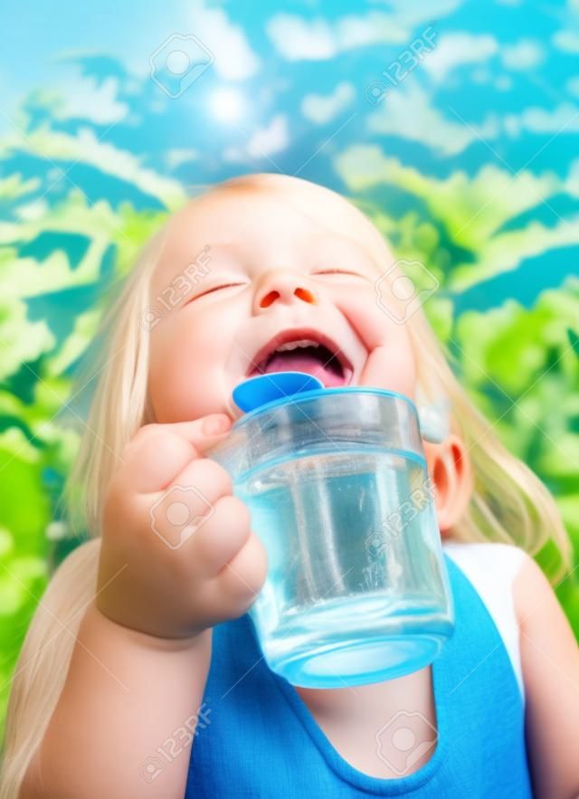 Blonde little girl wearing blue dress holding cup of fresh water with her eyes closed in a summer garden