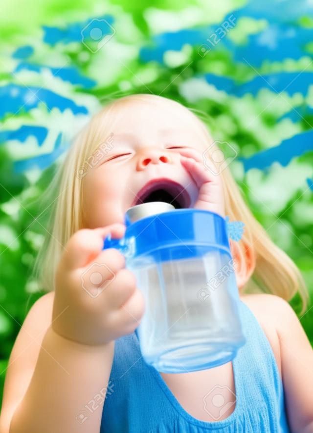 Blonde little girl wearing blue dress holding cup of fresh water with her eyes closed in a summer garden