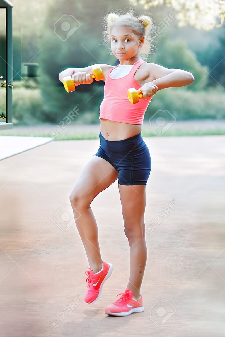 A cute, pretty teenage girl holds dumbbells in her arms and performs various outdoor exercises. Lifestyle