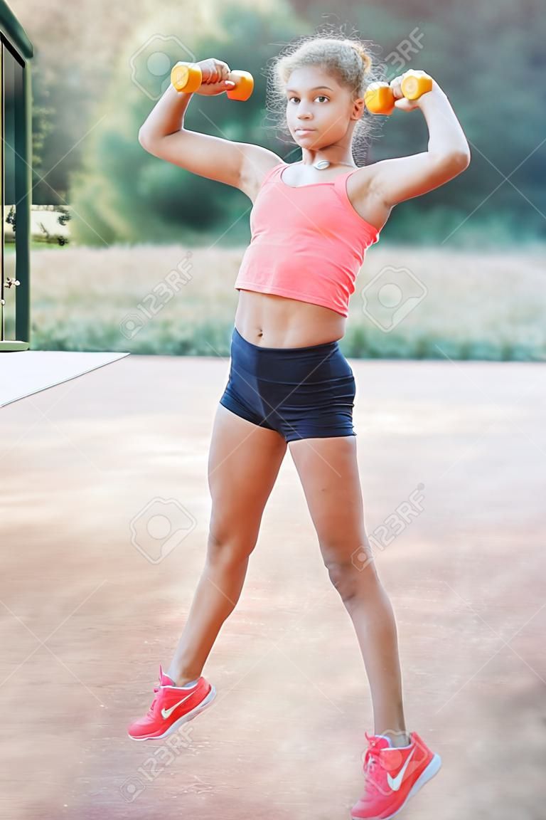 Una linda y bonita adolescente sostiene pesas en sus brazos y realiza varios ejercicios al aire libre. Estilo de vida