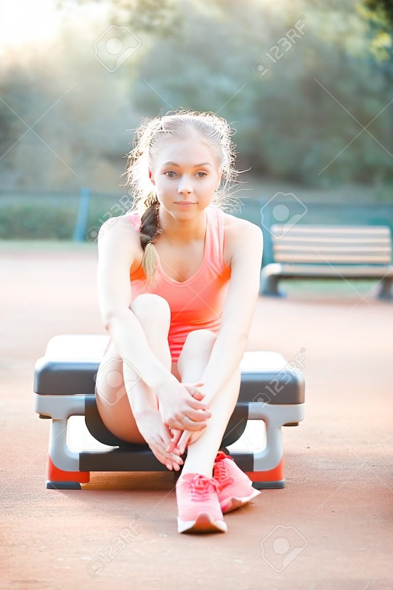 かわいい、かわいい十代の少女はステッププラットフォームに座って、屋外で彼女のトレーニングの後にリラックス