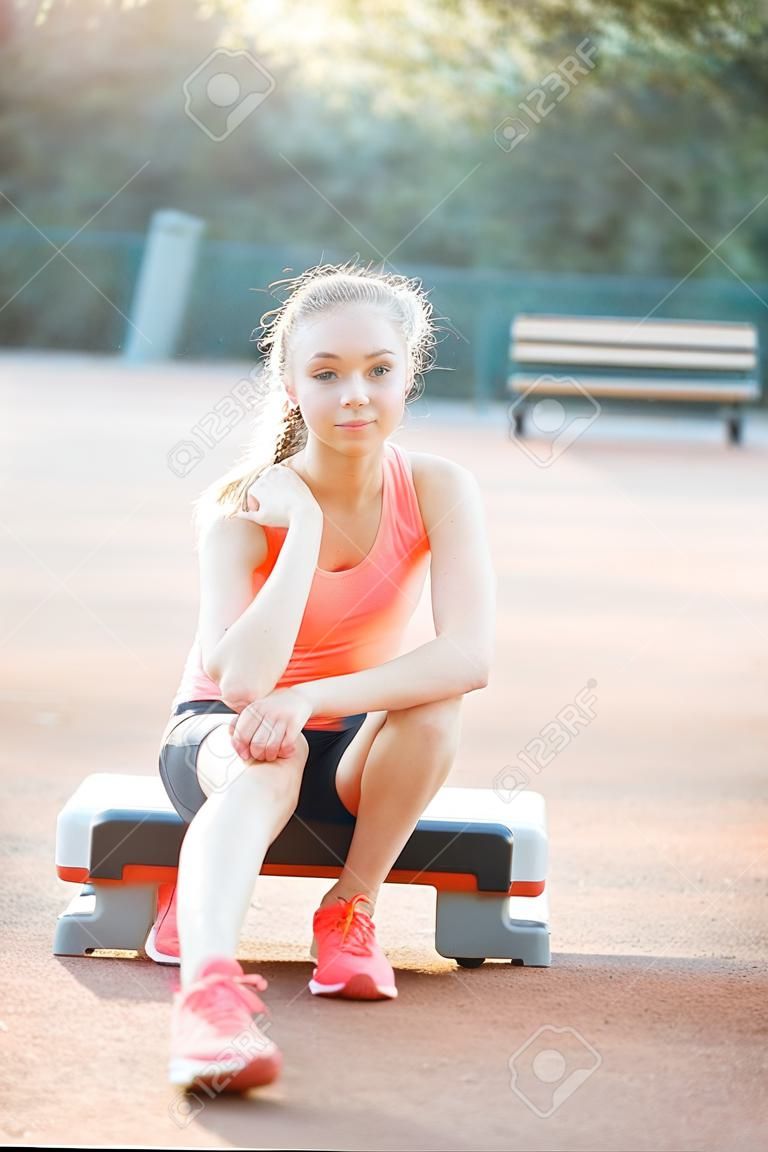 かわいい、かわいい十代の少女はステッププラットフォームに座って、屋外で彼女のトレーニングの後にリラックス