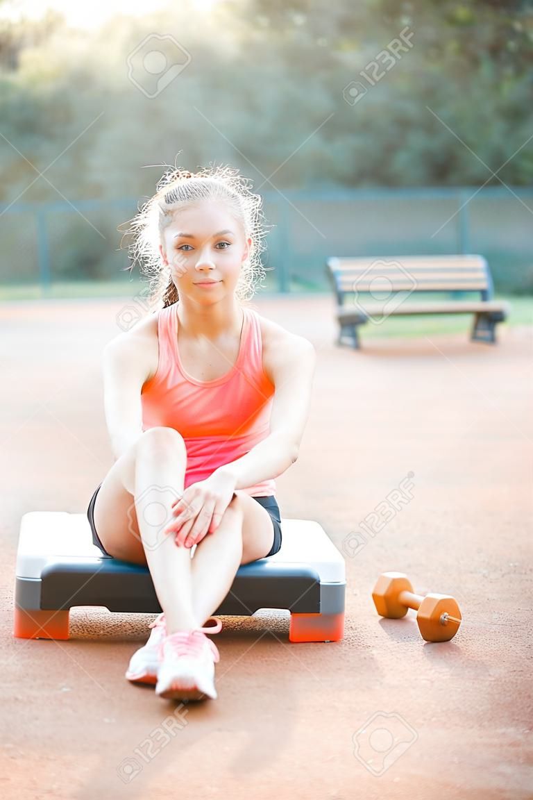 Uma linda e bonita adolescente senta-se em uma plataforma de passo e relaxa após seu treino ao ar livre